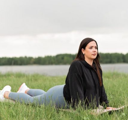 Woman lying on a yoga mat in a grassy field by a lake, promoting relaxation and wellness.