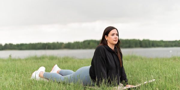 Woman lying on a yoga mat in a grassy field by a lake, promoting relaxation and wellness.